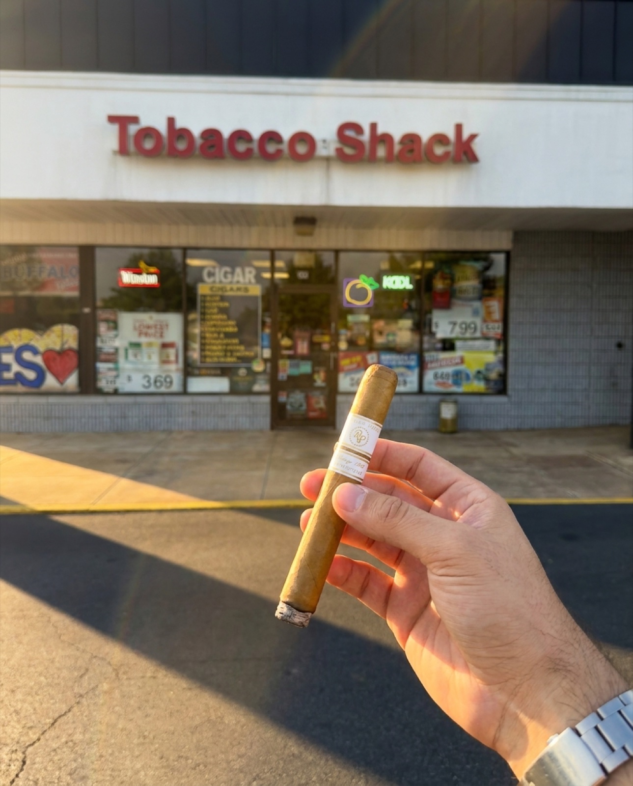A hand holding a cigar in front of the Tobacco Shack storefront at golden hour